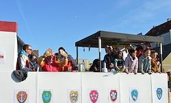 Eine Gruppe von Menschen, einige mit Jacken bekleidet, befinden sich auf einem Festwagen mit Schilden und einem Vordach. Sie schauen auf etwas vor ihnen.