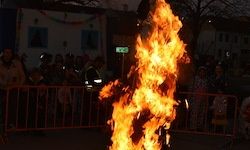 Eine Menschenmenge beobachtet eine Feuershow mitten auf einer Straße bei Nacht. Das Feuer ist hoch und hell, umgeben von einer Metallbarriere. Im Hintergrund befinden sich Gebäude mit Fenstern und Dekorationen.