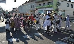 Eine Gruppe von Menschen in Kostümen, darunter weiße Bären, marschiert mit Musikinstrumenten die Straße entlang. Gebäude säumen die Straße, und Schatten werden auf den Weg geworfen.
