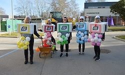 Fünf Personen, die in bunten Boxen mit Zahlen und Buchstaben gekleidet sind, stehen auf einem Parkplatz und halten Luftballons. Eine Person schiebt einen Wagen mit weiteren Luftballons. Gebäude und Bäume sind im Hintergrund.