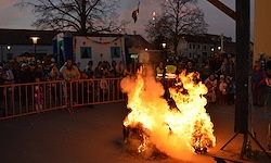 Ein Lagerfeuer brennt mitten auf der Straße, während Menschen zusehen. Ein Sicherheitsbeauftragter steht neben dem Feuer. Gebäude und Bäume sind im Hintergrund.