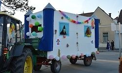 Ein dekorierter Traktor mit einem Anhänger in Form eines kleinen Hauses fährt auf der Straße, mit bunten Ballons und Dekorationen darauf. Im Hintergrund befinden sich Gebäude und Menschen.