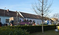 Ein festlicher Lastwagen mit Menschen oben und Bannern steht vor einem Haus. Ein Baum und Büsche sind in der Nähe, und andere Häuser sind im Hintergrund sichtbar.