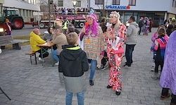 Eine Gruppe von Menschen in bunten Kostümen geht auf einer Kopfsteinpflasterstraße, ein Kind in einer Kapuzenjacke sieht sie an. Es gibt Tische mit sitzenden Leuten, und ein Traktor und ein Gebäude sind im Hintergrund.