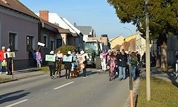 Ein Straßenumzug mit Menschen, die Schilder und Kostüme halten, einem Traktor im Hintergrund und Gebäuden auf beiden Seiten.
