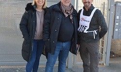 Drei Personen stehen vor einem Zaun, zwei Männer und eine Frau. Alle tragen Winterkleidung. Ein Mann hat eine weiße Schürze mit der Nummer 112.