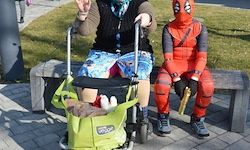 Eine Person in einem Deadpool-Kostüm sitzt auf einer Bank neben einer Person im Rollstuhl. Beide lächeln und posieren für ein Foto.