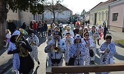 Eine Gruppe von Menschen in weißen Pünktchen-Kostümen geht die Straße entlang und spielt verschiedene Instrumente. Gebäude und Bäume im Hintergrund.