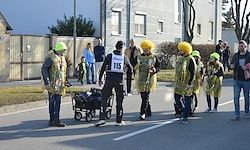 Eine Gruppe von Menschen in gelben Kostümen steht auf der Straße, einer schiebt einen Kinderwagen. Einige Personen tragen gelbe Mützen und Turnschuhe. In der Ferne befinden sich Gebäude und Personen in der Nähe eines Zauns.