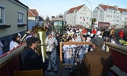 Eine Gruppe von Musikern in weißen und schwarzen Kostümen spielt Instrumente bei einem Straßenumzug. Die Menge schaut von der Straße aus zu, und Gebäude säumen die Straße.