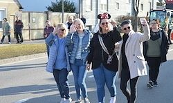 Eine Gruppe von Menschen geht eine Straße entlang, winkt und macht Fotos. Einige tragen Minnie-Maus-Ohren. Im Hintergrund sind Gebäude und ein Traktor zu sehen.