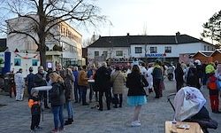 Eine Menschenmenge, darunter Familien mit Kindern, versammelt sich auf einem öffentlichen Platz. Die Umgebung ist von Gebäuden mit festlichen Dekorationen umgeben, und einige Menschen tragen Kostüme.