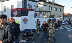 Ein Umzug mit einem großen Fahrzeug in Feuerwehruniformen, Leuten in Kostümen und Zuschauern, die vor Gebäuden auf der Straße gehen.