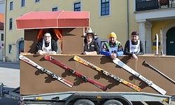 Vier Männer posieren in einem dekorierten Anhänger mit Skiern an einem sonnigen Tag. Sie lächeln und lehnen sich gegen den Anhänger. Dahinter befinden sich Gebäude mit Fenstern.