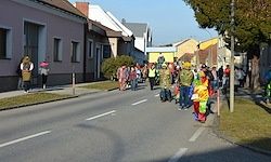 Eine Gruppe von Menschen in bunten Kostümen geht eine Straße entlang, umgeben von Gebäuden und Bäumen.