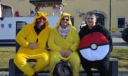 Drei Personen in gelben Kostümen und eine mit einem Pokémon-Ball sitzen auf einer Bank vor einem gelben Gebäude.
