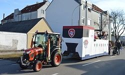 Ein roter Traktor zieht einen dekorierten Festwagen die Straße hinunter, mit Menschen im Inneren, vorbei an Gebäuden und einem Fußgänger.