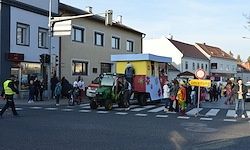 Ein dekorierter Dreirad-Traktor überquert eine Kreuzung in einer Stadt. Viele Menschen sind um ihn herum versammelt, wahrscheinlich für eine Parade oder Feier. Die Szene umfasst Gebäude, Fußgänger und Ampeln.