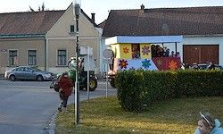 Ein dekorierter Festwagen mit Menschen wird von einem Traktor auf einer Straße gezogen. Eine Person läuft auf dem Bürgersteig. Mehrere Gebäude und geparkte Autos sind im Hintergrund.