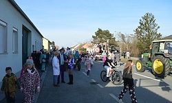 Eine Menschenmenge, einige in Kostümen, steht und läuft auf einer Straße. Ein Fahrrad und ein Traktor sind vorhanden. Gebäude und Bäume sind im Hintergrund.