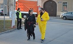 Ein Mann und eine Frau in Kostümen gehen die Straße entlang. Ein Polizeibeamter steht in der Nähe, und Fahrzeuge sind am Straßenrand geparkt.