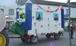 Ein mit bunten Ballons und Fenstern verzierter Anhänger, der von einem Traktor gezogen wird, bewegt sich die Straße hinunter. Im Hintergrund sind Gebäude mit Schildern zu sehen.