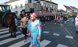 Ein bunter Umzug findet auf der Straße statt. Ein Mann in einem rosa Hut und blauem Kostüm geht. Dahinter fährt ein Lastwagen mit Leuten oben vorbei. Mehrere Menschen gehen auf der Straße und einige überqueren die Straße.