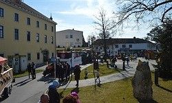 Eine Straßenszene mit Menschen, die auf dem Bürgersteig gehen, einem Bus und Gebäuden mit Bäumen und einem Steinmonument im Vordergrund.