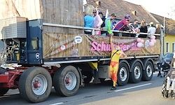 Ein dekorierter Lastwagen mit Leuten darauf, eine Person mit erhobenem Arm und einem Banner mit dem Text 'Schuster-Verni'.