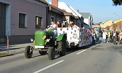 Ein grüner Traktor mit einem dekorierten Anhänger fährt die Straße entlang, mit Menschen, die darauf fahren, und anderen, die an der Seite zusehen.