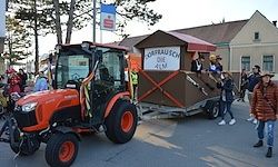 Ein roter Traktor zieht eine braune Kiste auf Rädern, in der Menschen sitzen, vor einer Menschenmenge und Gebäuden.