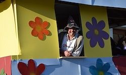 Ein Mann mit Hut und weißem Hemd sitzt in einer bunten hausartigen Struktur mit Blumendekorationen an den Wänden. Er lächelt und posiert für ein Foto.