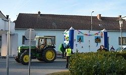 Ein grüner Traktor mit gelben Rädern fährt an einem dekorierten Haus vorbei. Eine Person in einer gelben Weste steht in der Nähe.