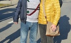 Zwei Personen stehen auf der Straße, ein Mann und eine Frau, beide tragen Taschen. Der Mann trägt eine blaue Jacke und Jeans, die Frau eine gelbe Jacke und schwarze Hose. Schatten fallen auf die Straße.