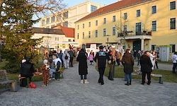 Eine Menschenmenge versammelt sich im Innenhof eines gelben Gebäudes mit vielen Fenstern, Bänken und einem Baum. Menschen in Winterkleidung gehen und sitzen auf Bänken.