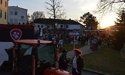 Sonnenuntergang über einer Stadt mit einem roten Traktor. Mehrere Menschen, einige in Kostümen, versammeln sich auf der Straße in der Nähe eines Gebäudes.