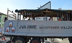 Ein Lastwagen mit 'JVA HME' darauf, hat ein vergittertes Dach mit mehreren Menschen darin. Er ist auf der Straße geparkt.