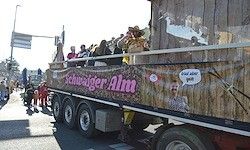 Ein Paradewagen mit einem großen Banner, auf dem 'Schweiger Alm' steht, hat Menschen darauf, die auf den Geländern stehen und zusehen. Es gibt Menschen, die am Straßenrand gehen.