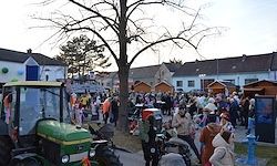 Eine Menschenmenge versammelt sich um einen grünen Traktor in einem Dorf. Ein Baum steht in der Mitte, mit mehreren Gebäuden und Straßenlaternen im Hintergrund.