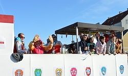 Eine Gruppe von Menschen in Kostümen steht auf einem geschmückten Wagen, winkt und lächelt in die Kamera. Der Wagen zeigt verschiedene farbenfrohe Embleme.