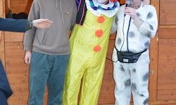 Drei Menschen in Kostümen stehen vor einem hölzernen Hintergrund. Ein Mann in einem Clownkostüm hält ein Mikrofon. Ein weiterer in einem grauen Hoodie und blauer Hose steht links.