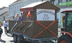 Ein Anhänger mit einer Holzkabine zeigt das Schild 'Dorfrausch die Alm'. Es sind Waffen an der Seite ausgestellt. Menschen gehen dahinter und ein Traktor ist rechts. Gebäude sind im Hintergrund.