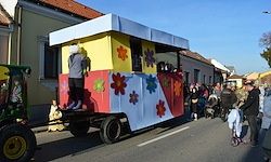 Ein farbenfroher Anhänger mit Blumen wird von einem Traktor auf der Straße gezogen. Menschen beobachten von der Straße.
