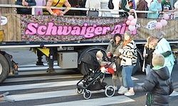 Ein Paradewagen mit einem rosa Banner mit der Aufschrift 'Schwaiger' steht auf einer Straße, mit Menschen und einem Kinderwagen im Vordergrund.