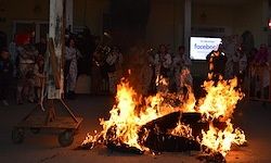 Ein Lagerfeuer mit einem großen brennenden Objekt in der Mitte, umgeben von einer Menschenmenge, einige in traditioneller Kleidung. Ein Wagen und ein Monitor mit der Aufschrift 'Facebook' sind ebenfalls zu sehen.