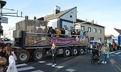 Ein großer Lkw mit einem geschmückten Anhänger fährt durch die Straße. Leute stehen herum, einige in Kostümen, und ein Banner mit Text ist am Lkw befestigt. Gebäude säumen die Straße.
