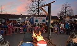 Eine Menschenmenge versammelt sich um ein Lagerfeuer, mit einer Person in einem rot-weißen Hut davor. In der Mitte der Menge steht ein Galgen.