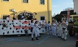 Eine Gruppe von Musikern in Kuhkostümen tritt auf einem geschmückten Wagen vor einem gelben Gebäude auf.