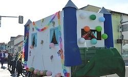 Ein dekorierter Festwagen mit bunten Luftballons, Girlanden und einer Person im Inneren, bei einer Straßenparade. Der Wagen ist in Weiß, Blau und Grün bemalt.