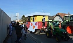 Ein bunter Umzug mit Menschen und Blumen wird von einem grünen Traktor an einem sonnigen Tag gezogen. Eine Person steht auf dem Aufsatz.